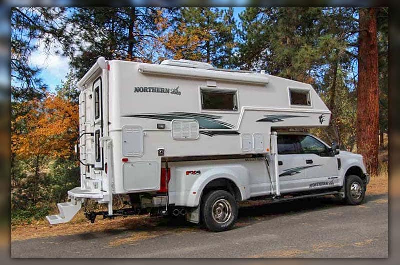 Northern Lite 10-2 EX CD SE truck camper parked in a picturesque camping spot surrounded by trees and mountains.