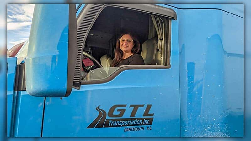 Young woman studying "Semi Truck Driving For Beginners" manual inside a semi truck cab.